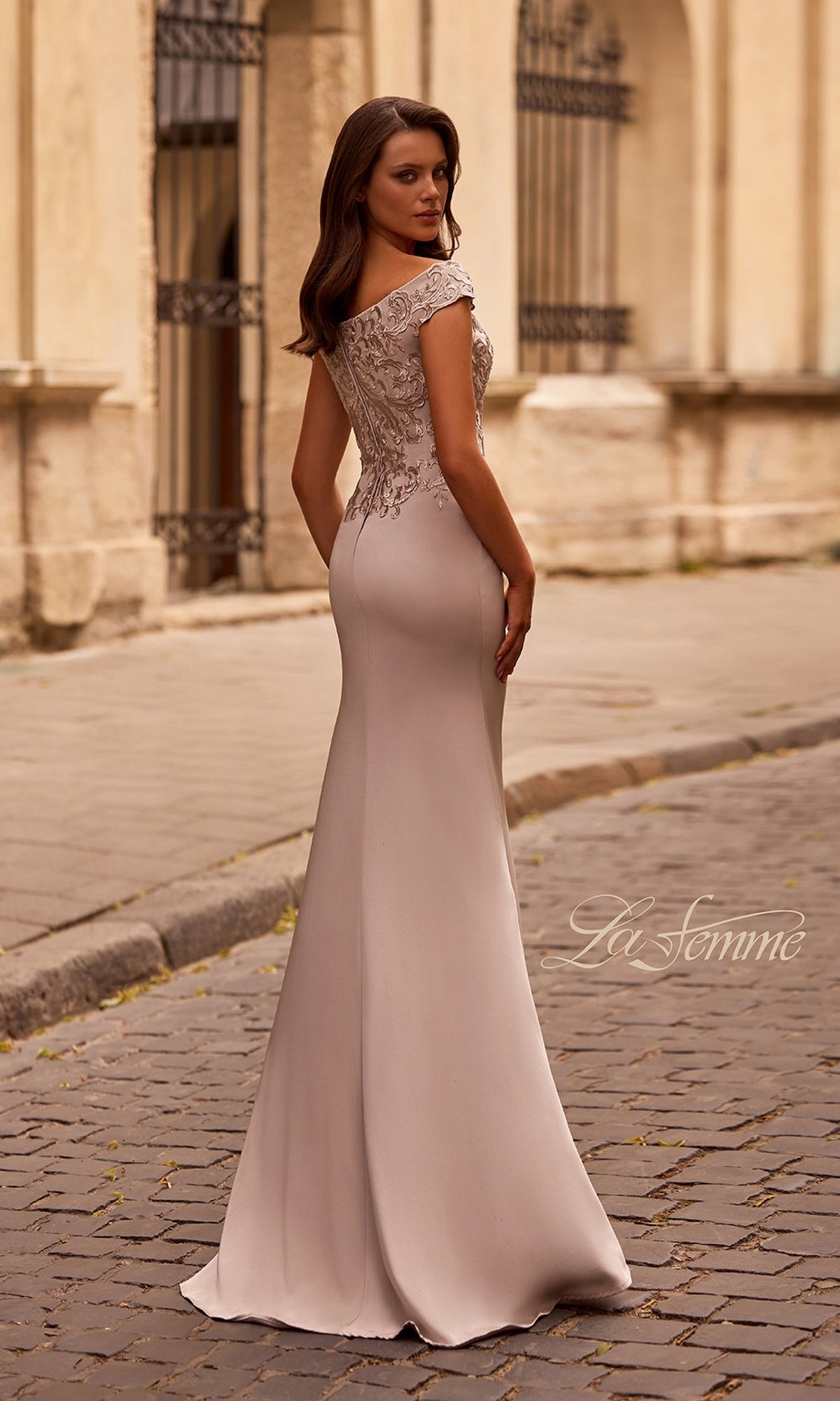 A woman in the Long Formal Dress: La Femme 29331, featuring a fitted embroidered bodice and off-the-shoulder neckline, stands on a cobblestone street looking back.